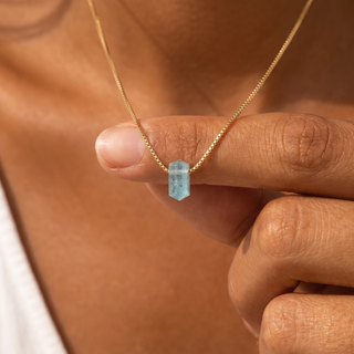 Eisblauer Aquamarin Zylinder zwischen zwei Fingern, Goldkette im Hintergrund