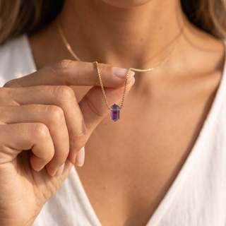 Frau hält den Amethyst Zylinder an der feinen Goldkette vor weißem Oberteil