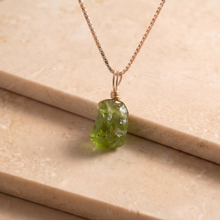 Nahaufnahme des grünen Peridot Rohsteins an Goldkette auf Travertin