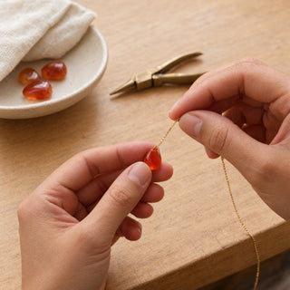 Hands threading a fine gold chain through a carnelian teardrop in the Handmade Art by Carola studio