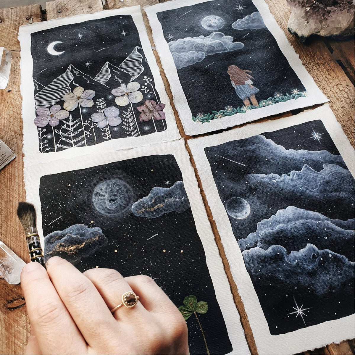 Four pictures painted by Carola with the night sky, moon and clouds, next to her brush