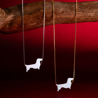 Zwei Dackelketten mit weißem Dackel in Silber und in Gold an einem Ast vor rotem Hintergrund