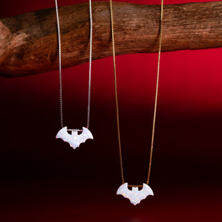 Zwei Fledermausketten mit weißer Fledermaus in Silber und in Gold an einem Ast vor rotem Hintergrund